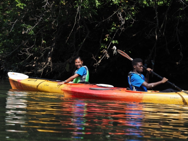 Two young people are smiling and paddling on bright kayaks.