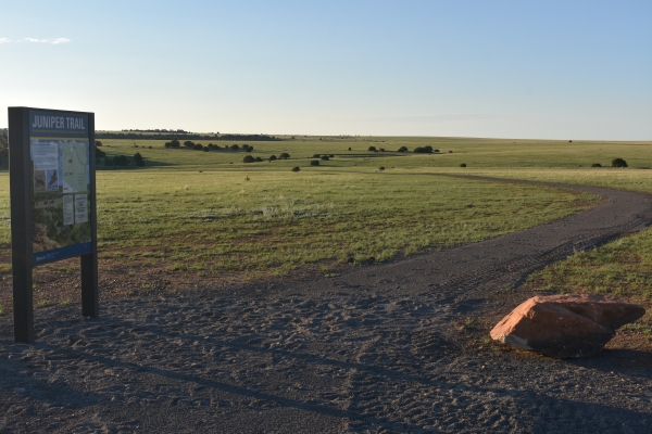Trailhead sign that says "Juniper Trail" at Rio Mora National Wildlife Refuge on left. Trail going off into the distance with a rock on the right. . 