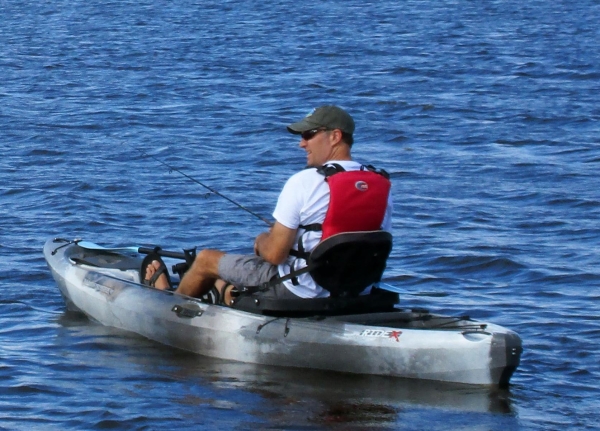 Man in white shirt and red PFD holding a fishing pole in a gray kayak surrounded by blue water.