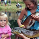 Two girls pose with two big trout in front of a pond where other children are fishing.
