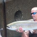 A man in waders and sunglasses holds up a large salmon horizontally with both hands.