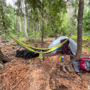 A campsite with a hammock hanging between two trees, a tent, boots, and other gear on the ground.