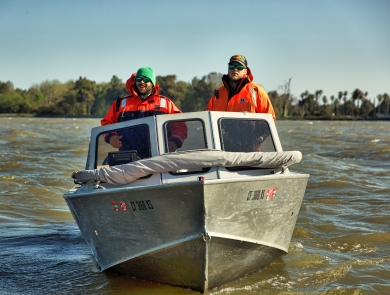 Two men pilot a motored boat on a river