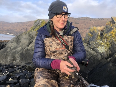 Biologist Robin Corcoran in chest waders and cold weather clothing holding a surfbird with both hands while sitting near a rocky shoreline in Alaska .