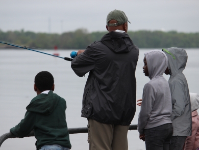 A man stands on the waterfront with a fishing rod surrounded by kids