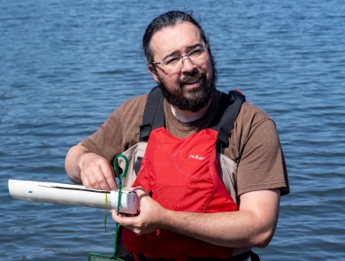 A service intern wearing waders and a PFD smiles as he measures the length of a fish. Water can be seen in the background.