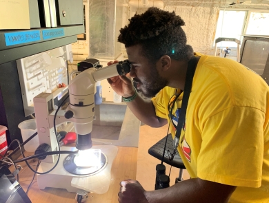 A young man in a bright yellow shirt leans in to look through a microscope in a lab. 