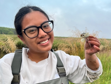 A Pathways employee smiles as she holds up a prickly sculpin. A field and sky can be seen in the background. 