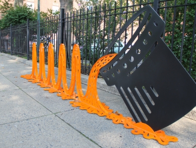 a metal art installation of orange waves pouring from a black can sits on a sidewalk, with a black metal fence and green shrubs in the background