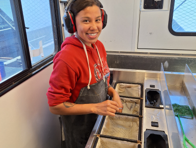 AmeriCorps intern in a fish marking trailer wearing a bib and ear protection. In front of her are four submerged net baskets, two containing juvenile coho salmon.