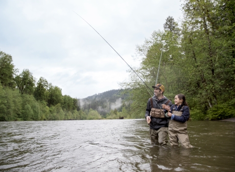 A parent and child stand in a river, fly fishing.