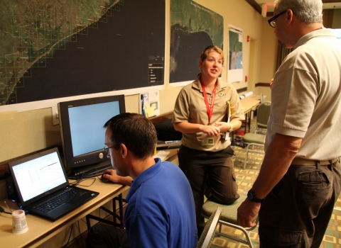 Three people are busy at work in the Operations Center during the aftermath of the Deepwater Horizon oil spill. One person is focused on a computer screen, with a map of the impacted area over their head. The two other people are talking to each other. 