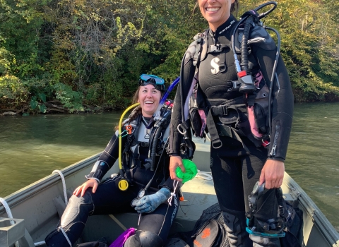 Two people wearing diving gear. One standing and one sitting in a boat and both smiling happily.