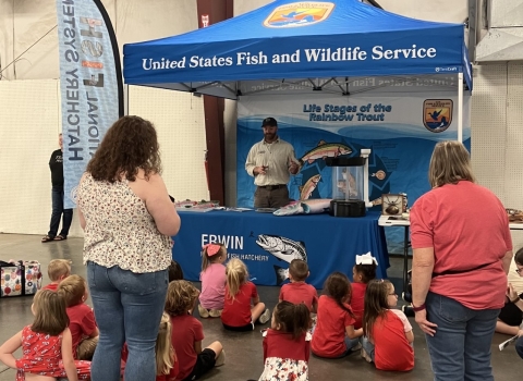 A group of people standing and sitting on the floor while listening to a presentation.