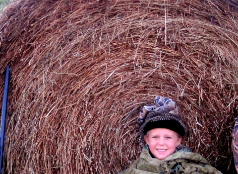 A mother holding a hunting rifle with her two young daughters crouching next to a bay of hale in a green field. All three are in camouflage.