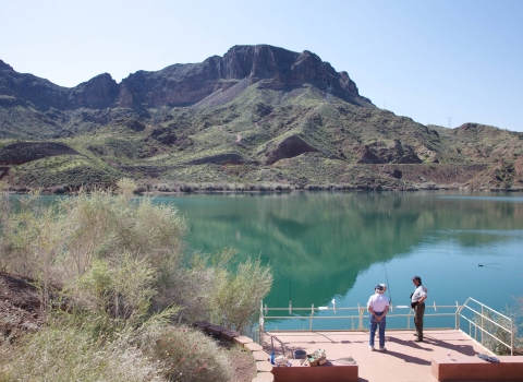 Two people fish at a pier with mountains in the background.