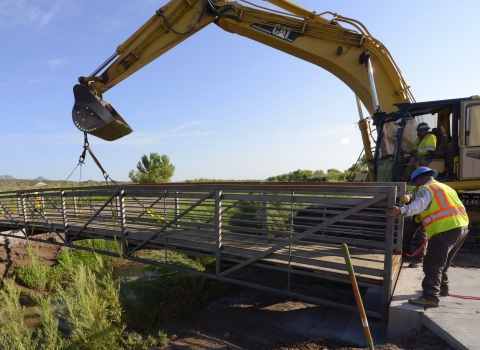 A crane operator lowers a bridge into place while a coworker on the ground adjusts the placement. 