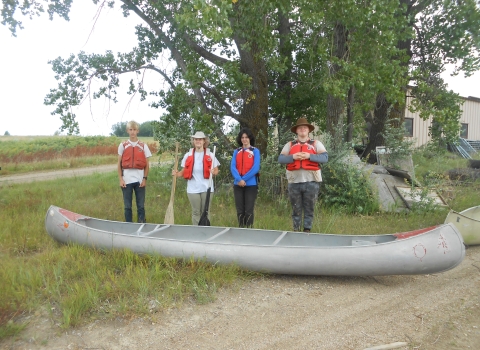 4 youth standing in a line behind an aluminum canoe outside
