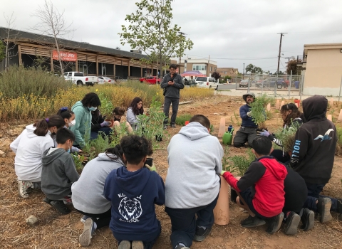 A group of students kneel next to each other in a semi circle as their teacher stands on the left end of the semi circle. Each student has a single potted plant in front of them. They are either looking at their plant or listening to their instructor who is also in a kneeling-sitting position holding out a green and shrubby potted plant.