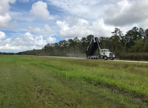 A dump truck tips a load of gravel onto a gravel road bordered in field and forest.