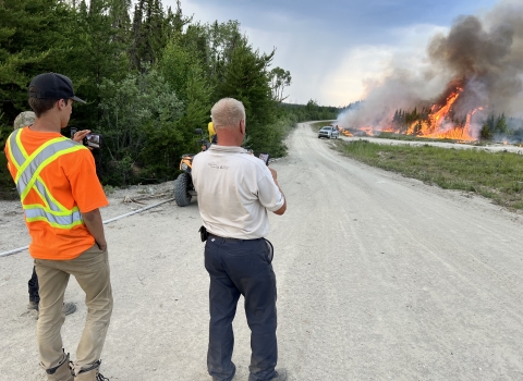 Three people watch flames burn through the forest with a truck near it along a dirt road