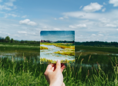 A hand holding up a tiny watercolor of the scene behind it