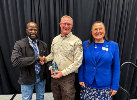 A unformed FWS employee shakes hands with another individual while holding an award. A woman stands smiling to the right of the awardee.