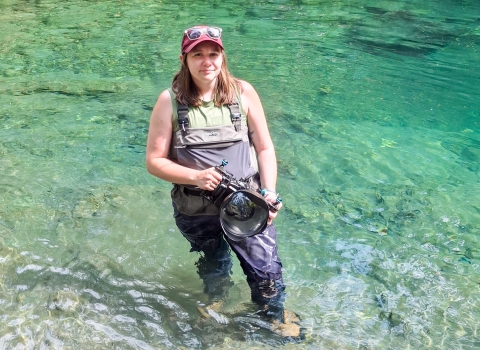 A service member wearing waders stands in a river holding a camera.