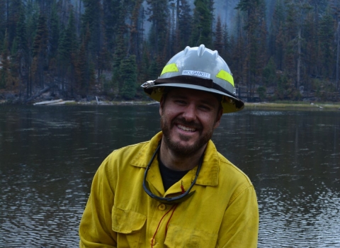 A firefighter dressed in Nomex protective clothing and hard hat, smiles and leans on their tool. Behind them is a lake and tall evergreen trees. The sky is hazy with smoke.