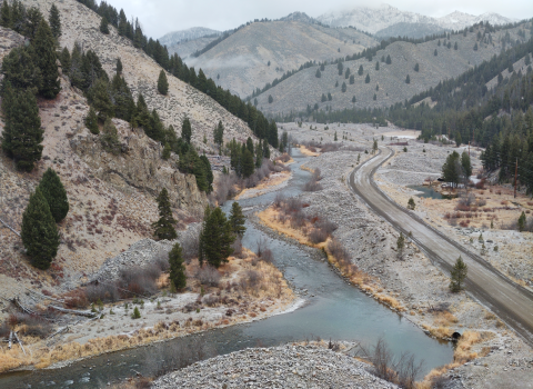 An aerial view over a winding river with a road along one side and mountains with evergreen trees on the other. there is a light dusting of snow on the road and terrain.
