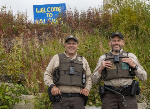 Two uniformed Federal Wildlife Officers pose for a picture with a painted sign in the background on a hill reading "welcome to Kalskag" 