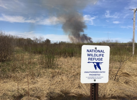White and blue sign in the foreground that reads National Wildlife Refuge Unauthorized Entry Prohibited with a blue goose in flight logo and spring native grasses and blue sky with a column of smoke in the distance.