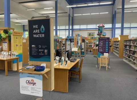 Photo of We Are Water MN Exhibit as displayed at North Branch Area Library as part of hosting experience with Chisago County. In the foreground is a welcome panel with the We Are Water Logo, as well as logos for many state partners. The background shows many other exhibit pieces at a distance, including a 12 foot tall water tower that is part of the exhibit. 