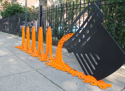 a metal art installation of orange waves pouring from a black can sits on a sidewalk, with a black metal fence and green shrubs in the background