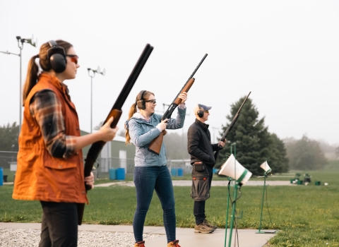 Three people wearing eye and ear protection stand with firearms at a skeet target range.