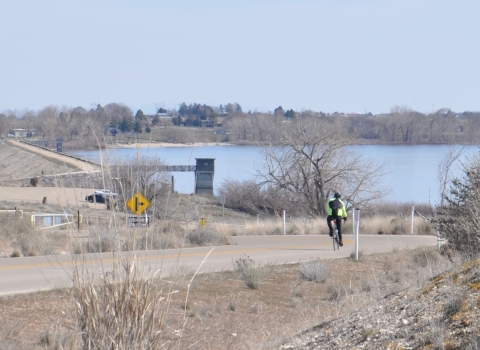 A person riding a bike on a narrow, two-lane road heading toward a lake and a dam.