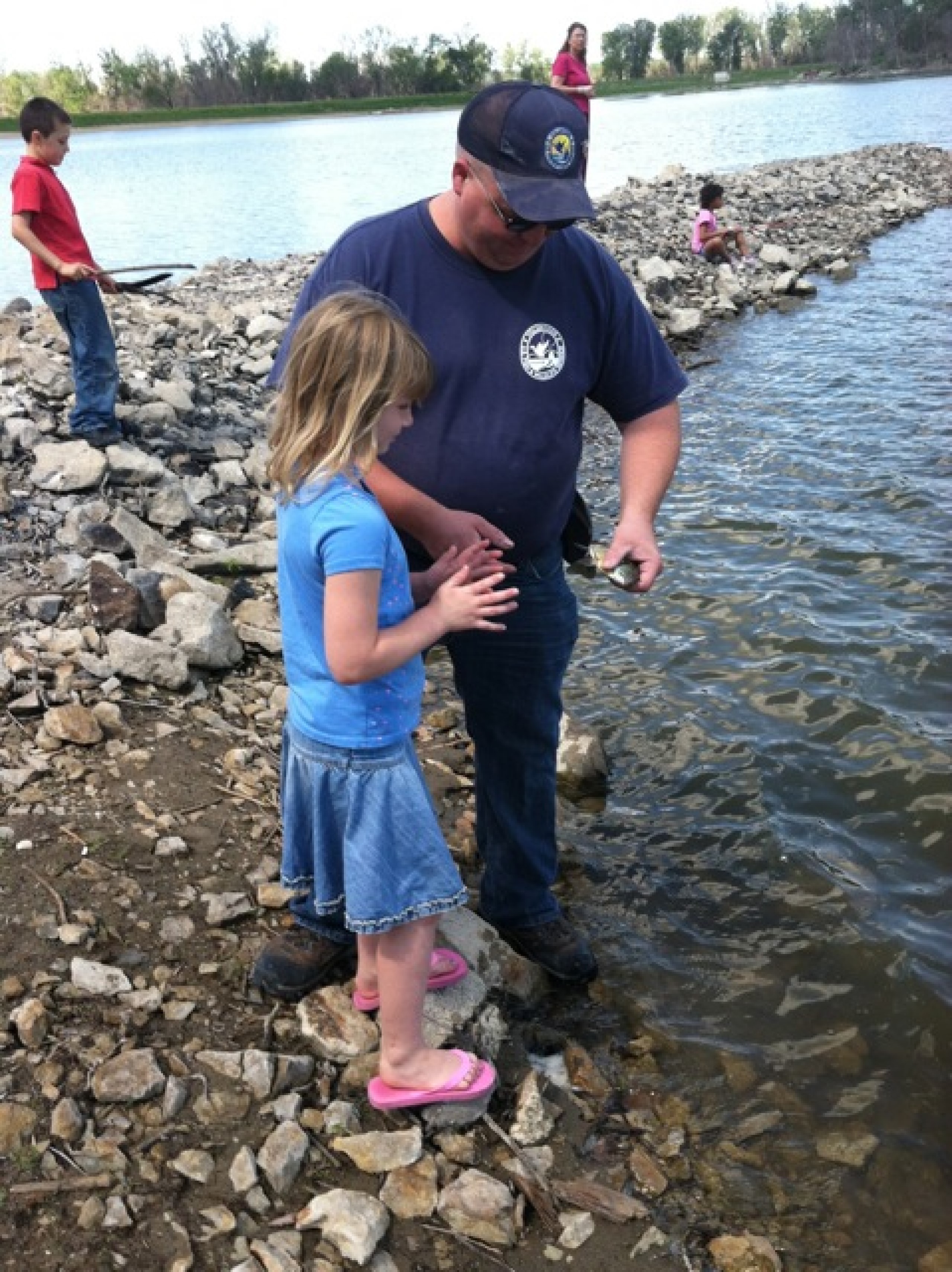 A refuge volunteer helping a young girl fix her fishing pole on a rocky shoreline.