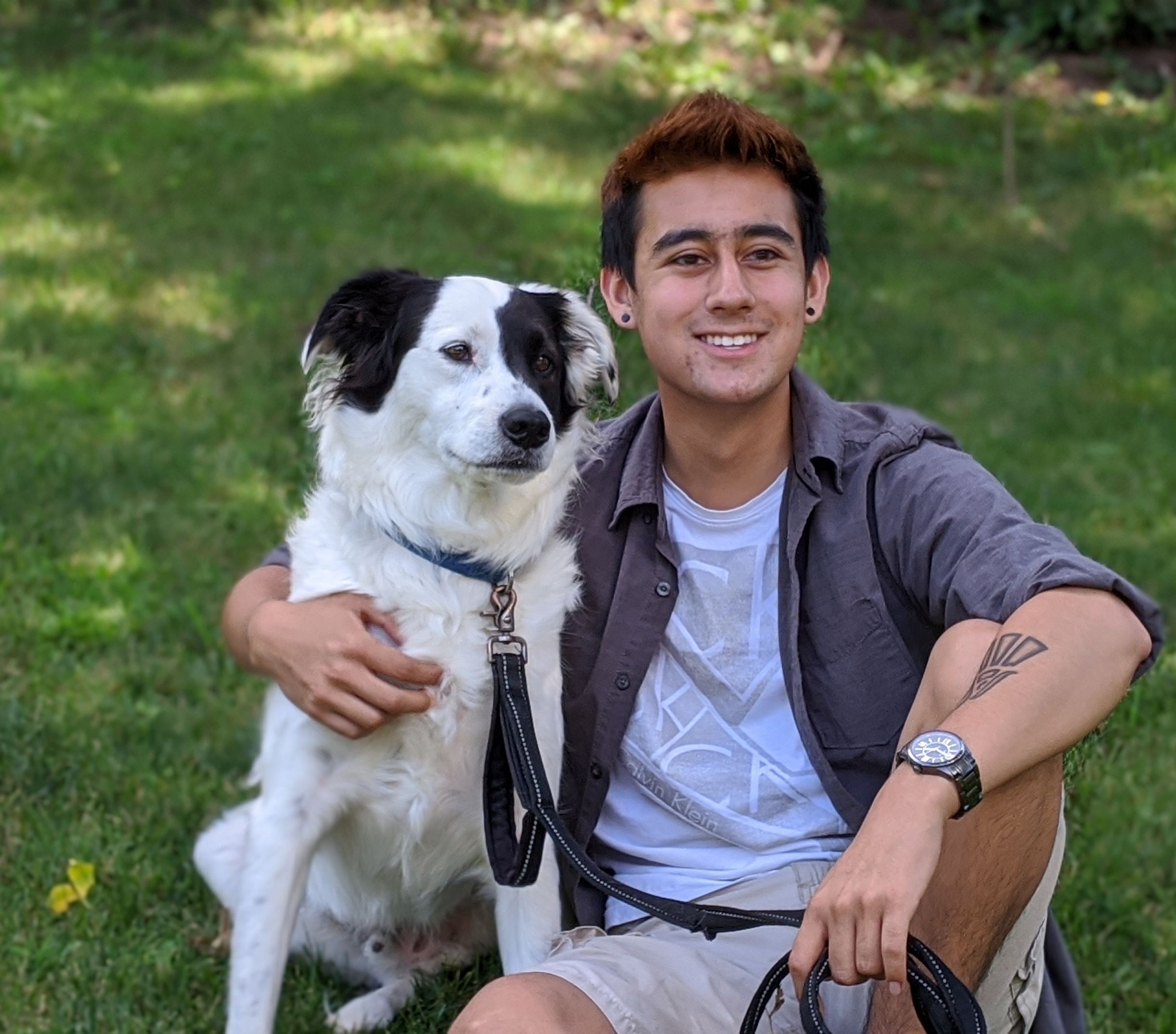 A smiling young man sits on the grass with his arm around a white and black dog.