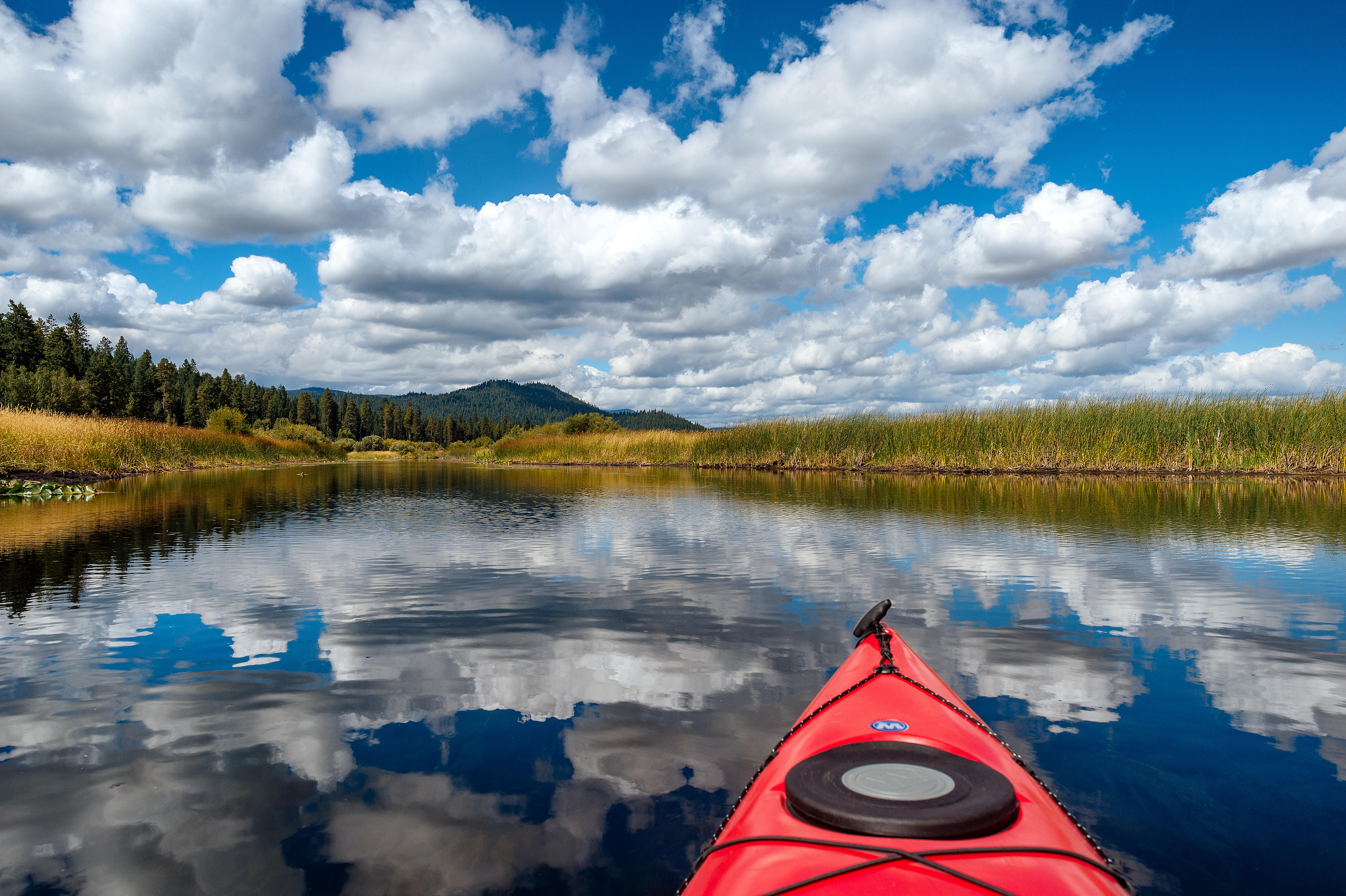 The front end of a red kayak points into clear water under a bright blue sky with some low clouds.