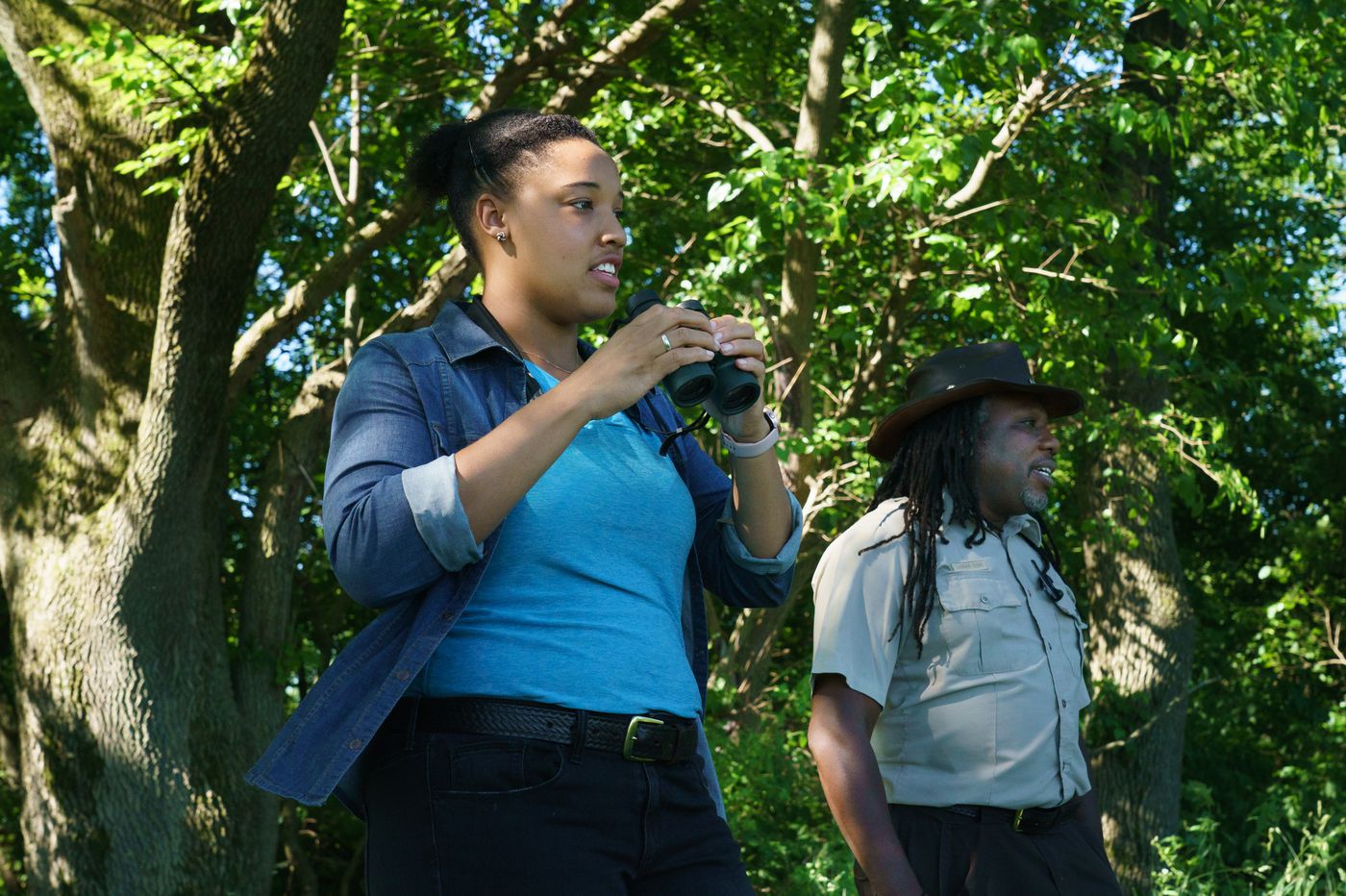 A woman and a man, both U.S. Fish and Wildlife Service employees, look for birds in the woods. The woman holds binoculars.