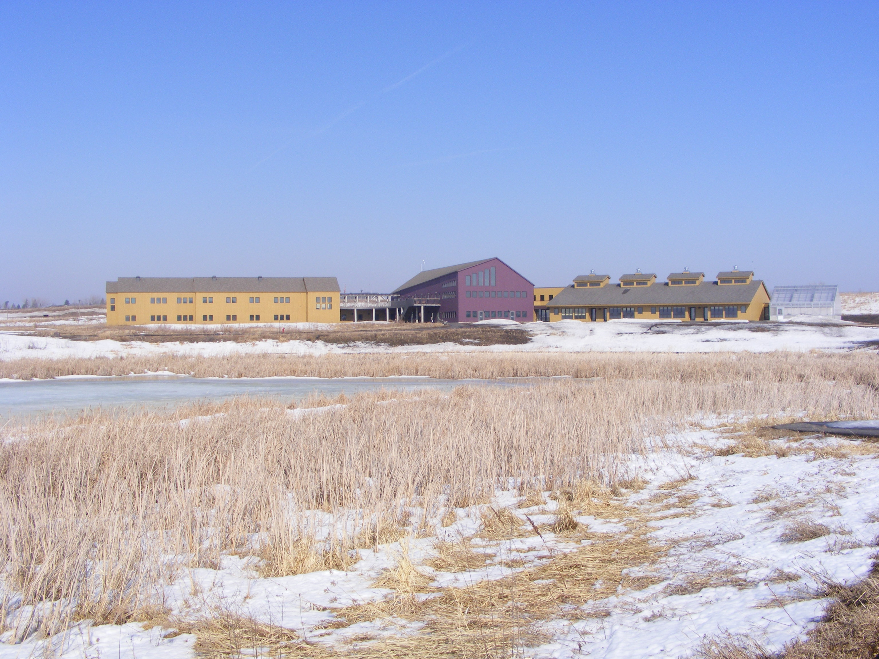 The Prairie Wetlands Learning Center buildings on the horizon below a sunny blue sky and above the snowy, tan prairie