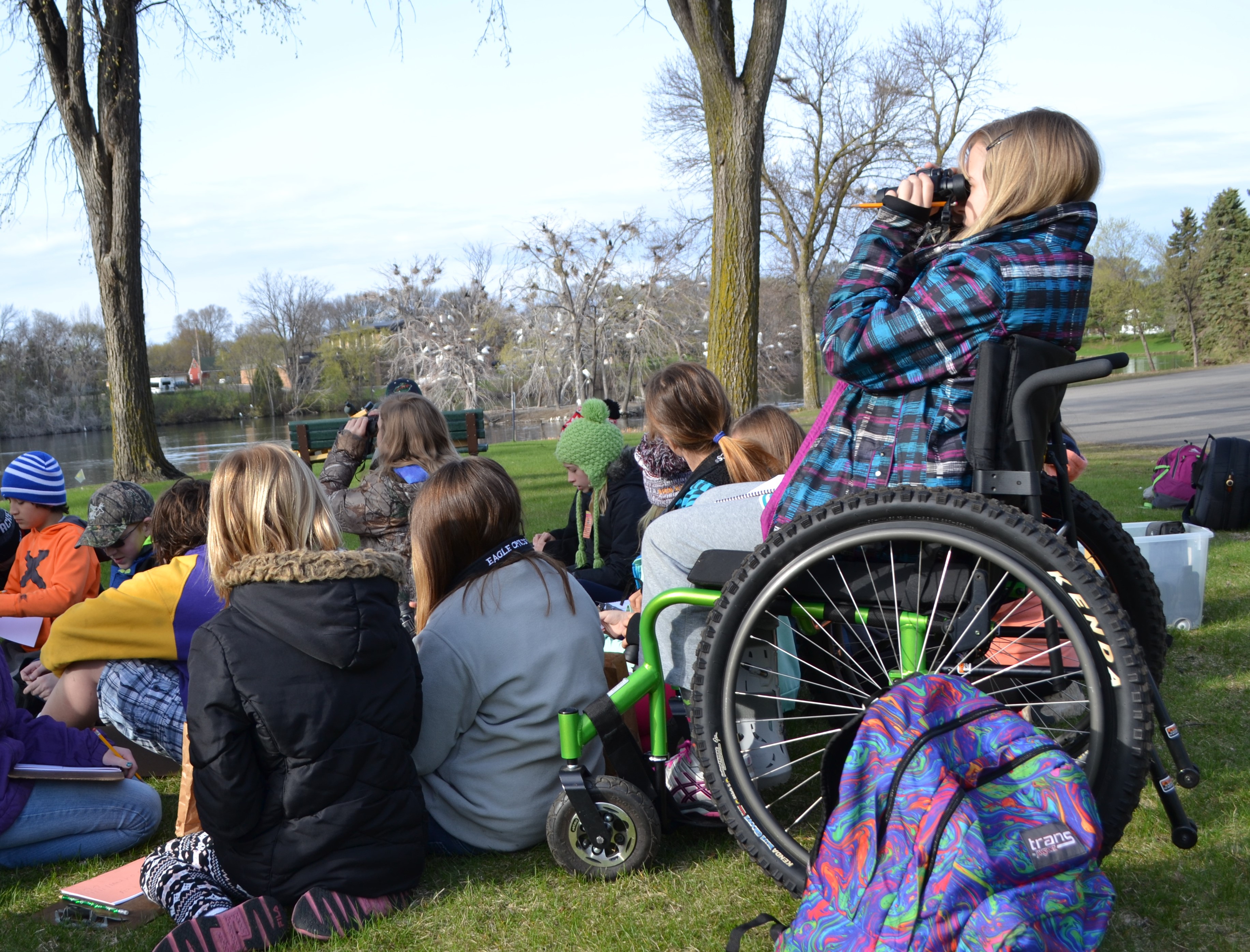 A student in a wheelchair using binoculars to watch birds with her class sitting on the ground