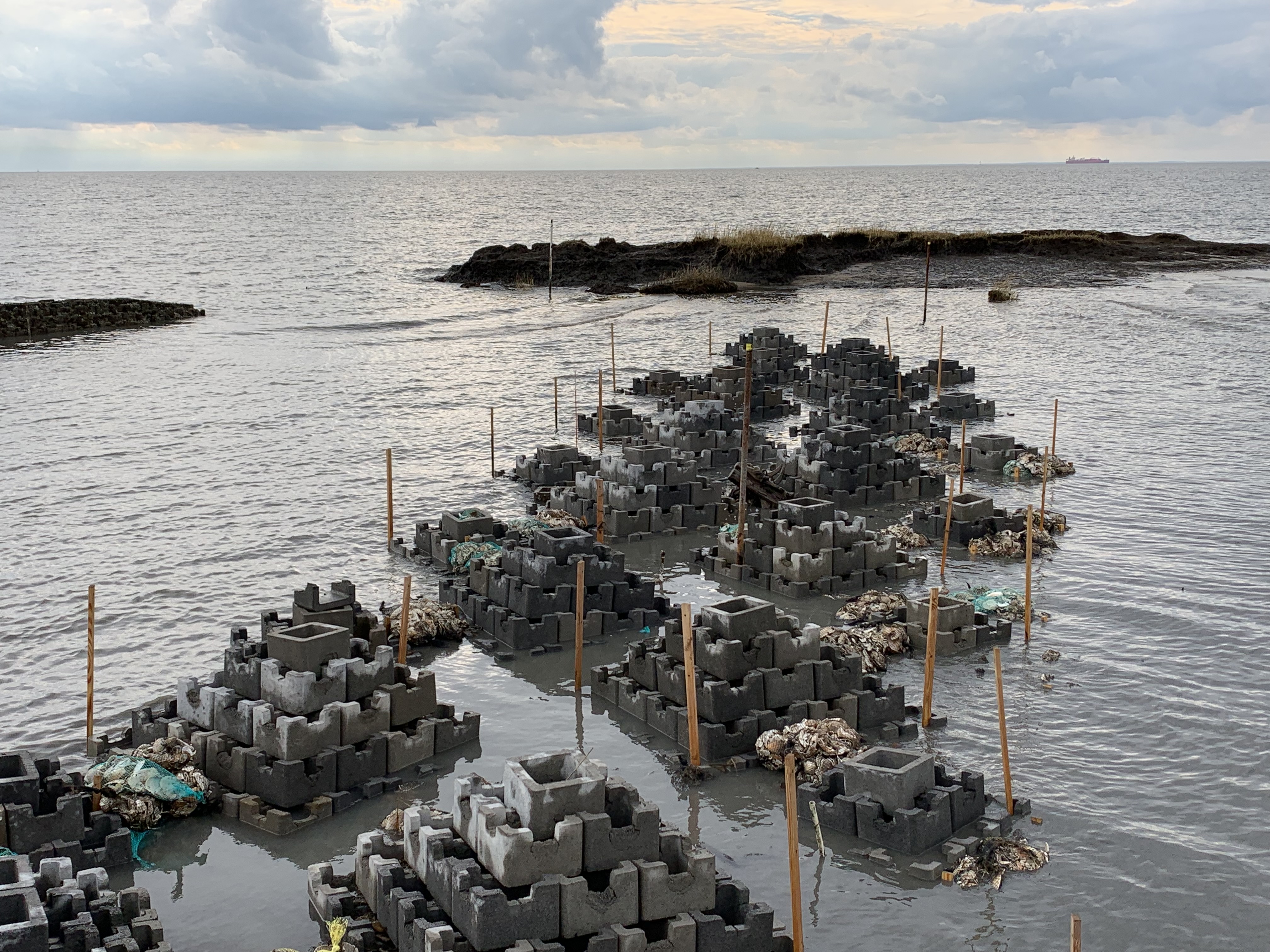 Several pyramid-type structures made of small concrete blocks sit in the water with an island-like peninsula behind them. 
