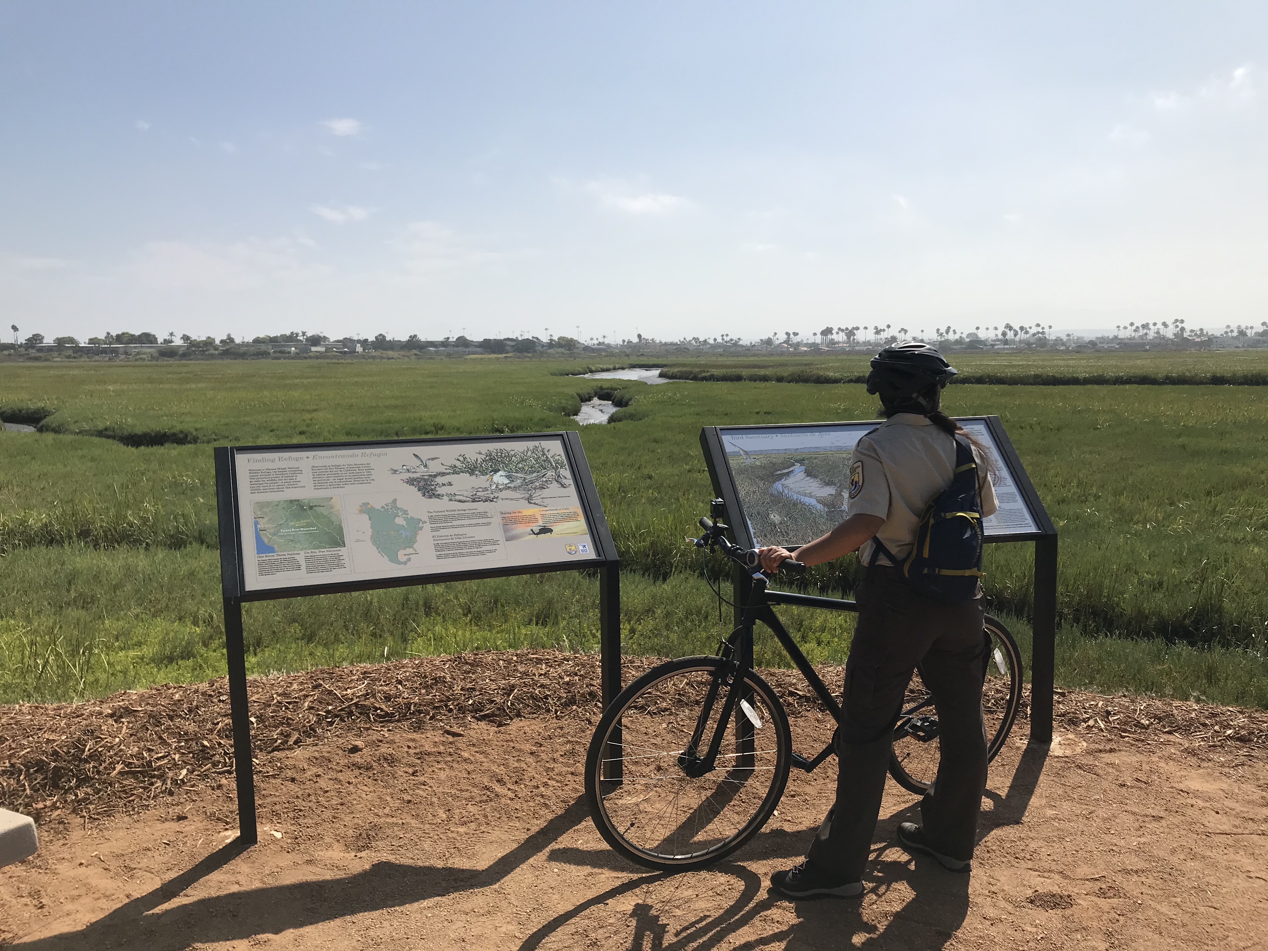 Interpretation panels near estuary. Park ranger holding bike in front of them reads the panel. 