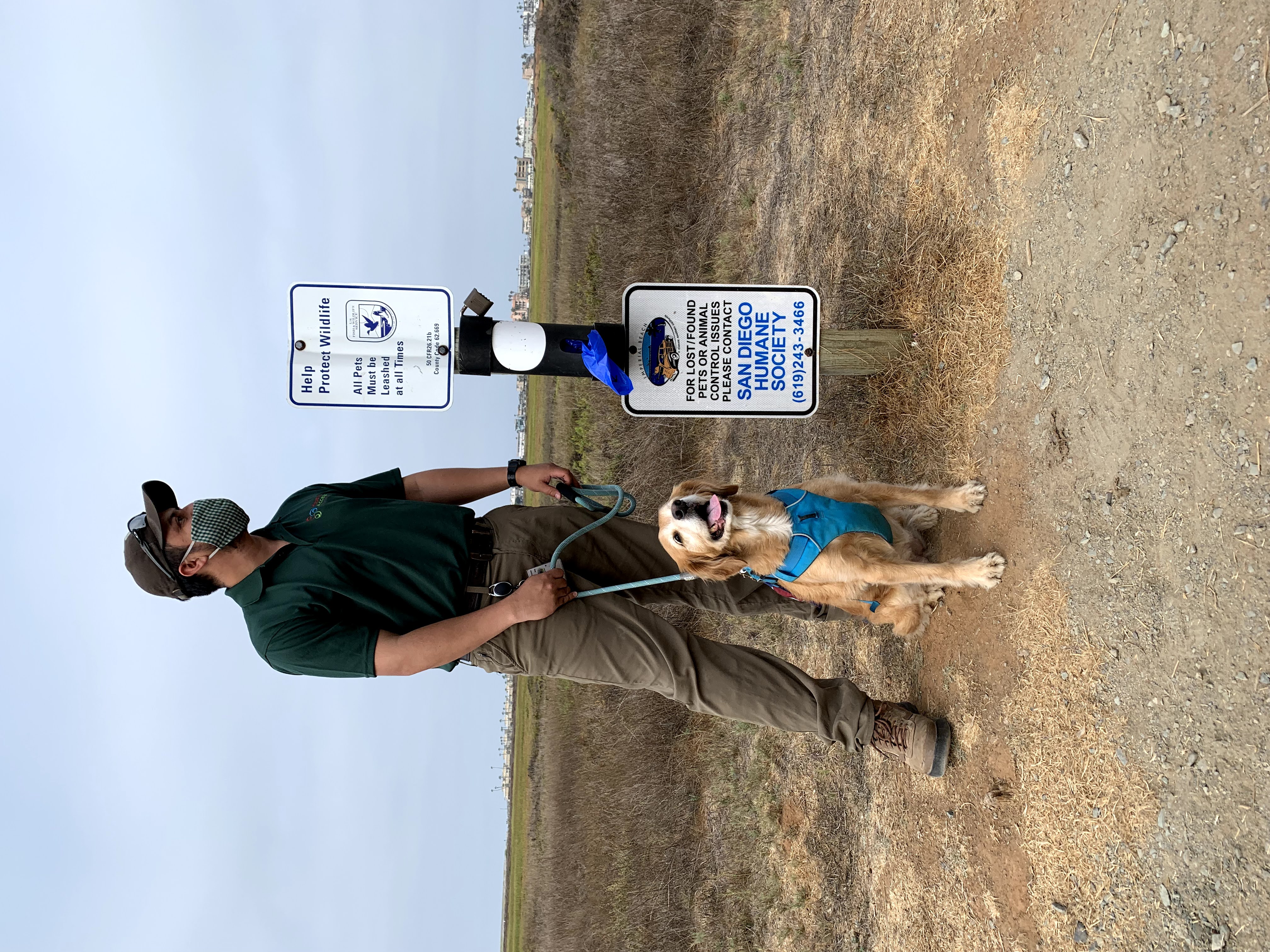 Individual wearing face mask with leashed dog near sign and waste bags.