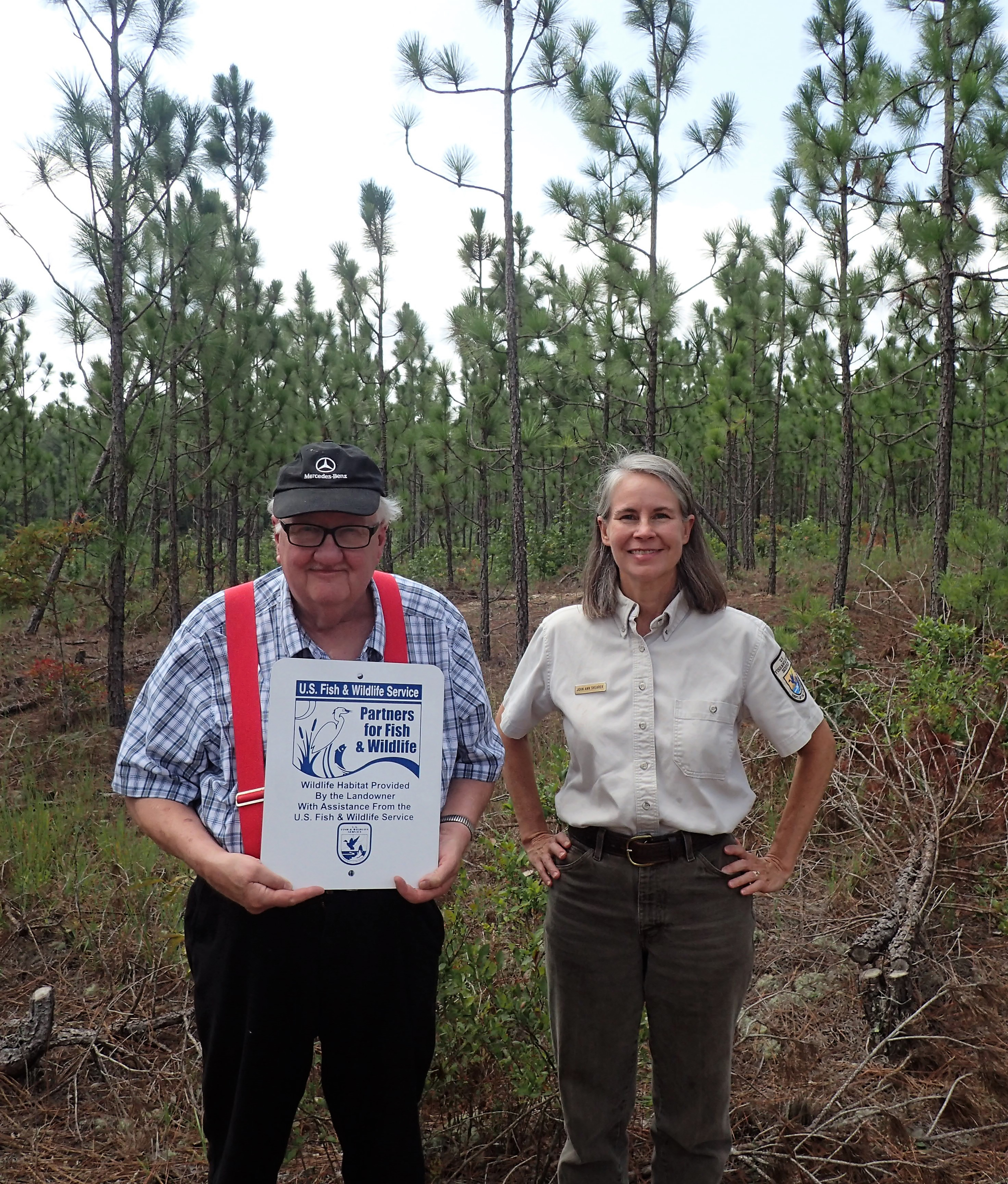 man holds a sign that reads "Partners for Fish and Wildlife". A woman in uniform stands next to him and they are both facing the camera with a young pine forest behind them.
