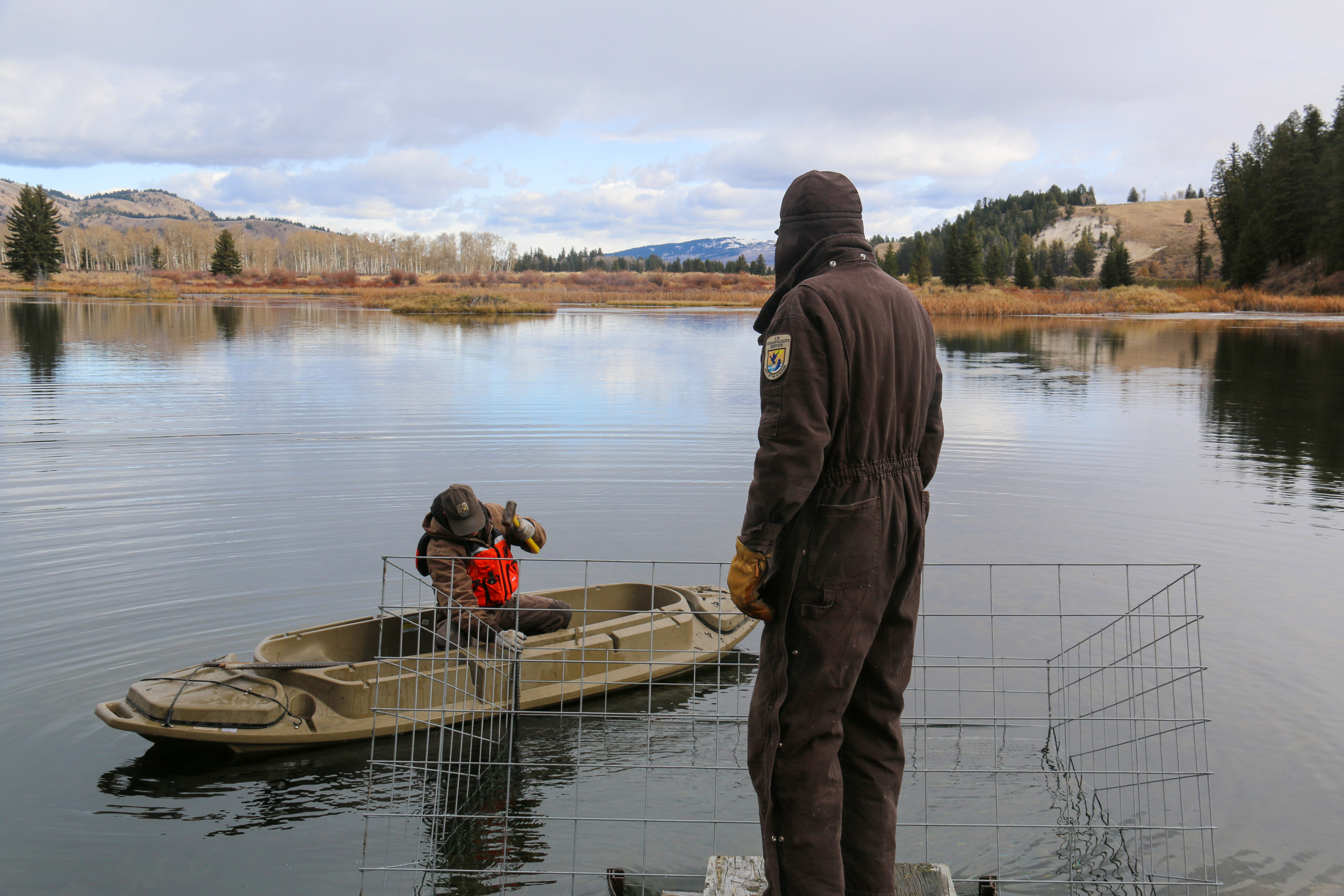 A biologist on a kayak in water installs a wire box while a USFWS employee watches from shore.