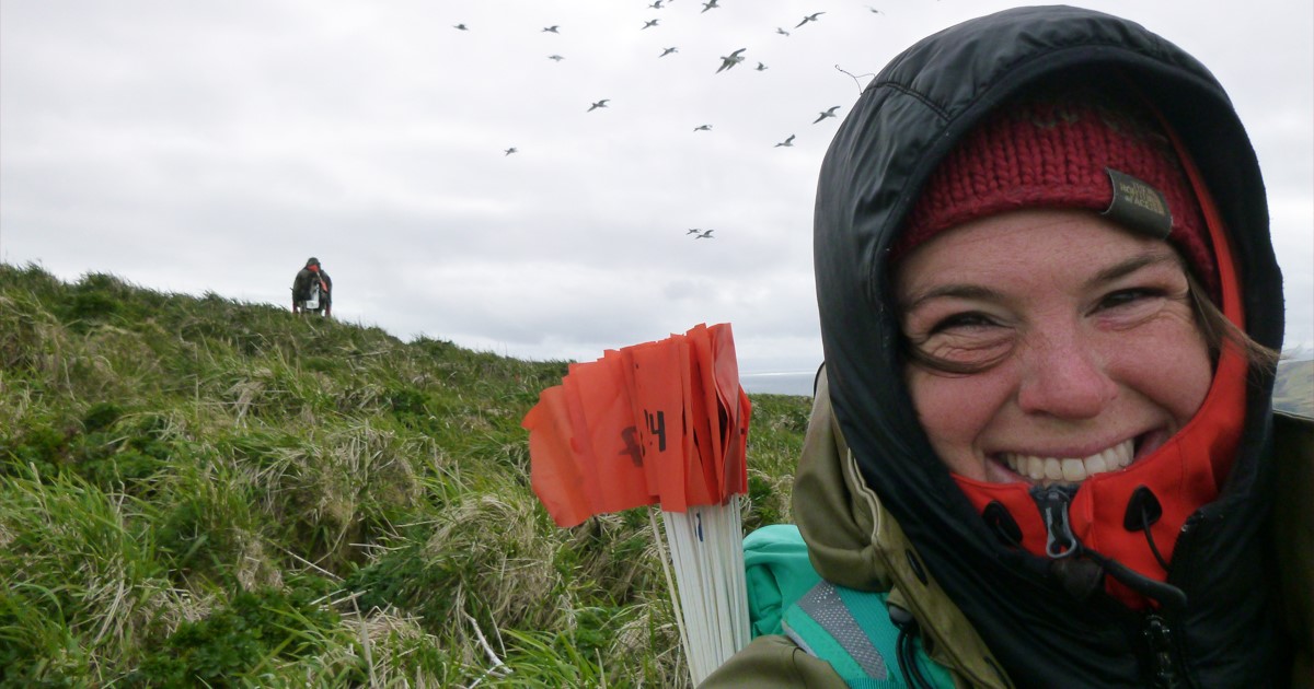 Smiling woman takes a selfie. She is wearing layers and a hood. There are survey flags, birds, a grassy hill, and another person over her shoulder and in the distance.