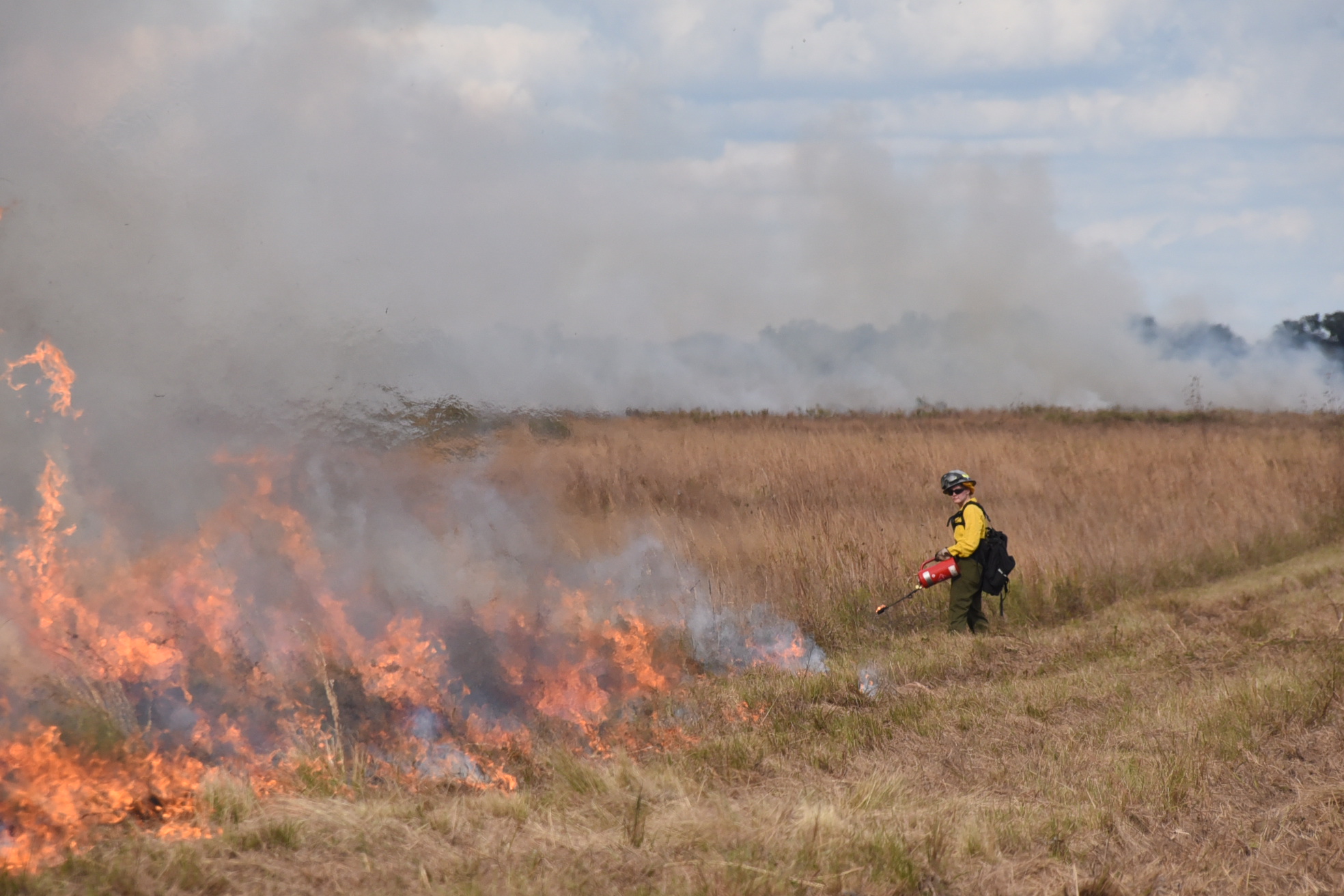 Fire burns prairie grasses as one of the crew carries a fuel torch to light. Smoke shrouds the horizon.