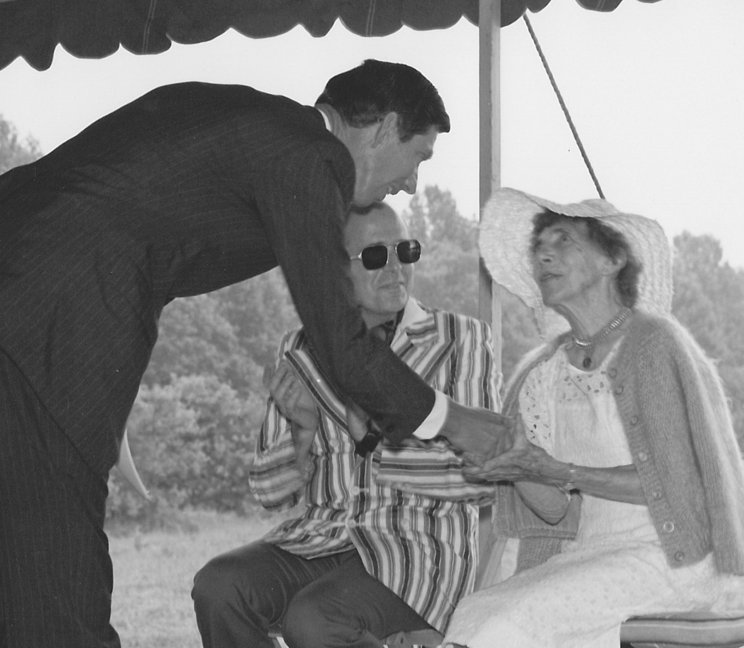 B&W photo of man in dark suit shaking hands with seated woman wearing a hat; man in striped sportcoat and sunglasses next to him